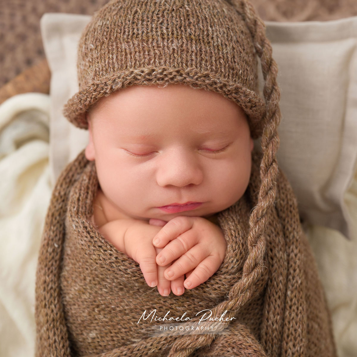 Nahaufnahme eines schlafenden Neugeborenen, eingewickelt in braune Strickstoffe und mit passender Strickmütze, die kleinen Hände sanft übereinandergelegt.